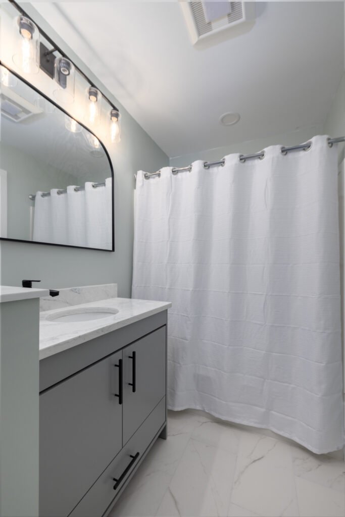 Modern bathroom remodel featuring a gray shaker vanity with a marble-look countertop, matte black hardware, an arched mirror, and marble-look tile flooring by GLG Homes.