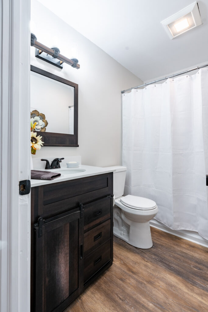 Modern bathroom remodel featuring a dark wood vanity with sliding barn door hardware, white countertop, black fixtures, wood-look flooring, and a tub-shower combination by GLG Homes.