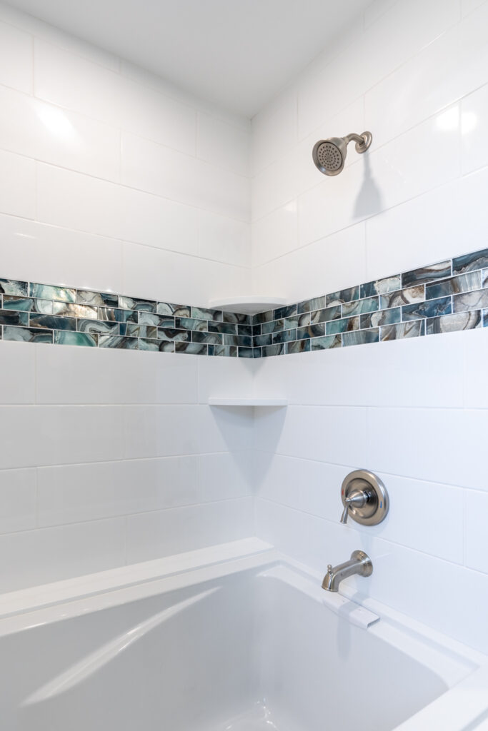 Custom bathroom shower with white subway tile, a blue and gray glass mosaic accent border, built-in corner shelves, and brushed nickel fixtures by GLG Homes.